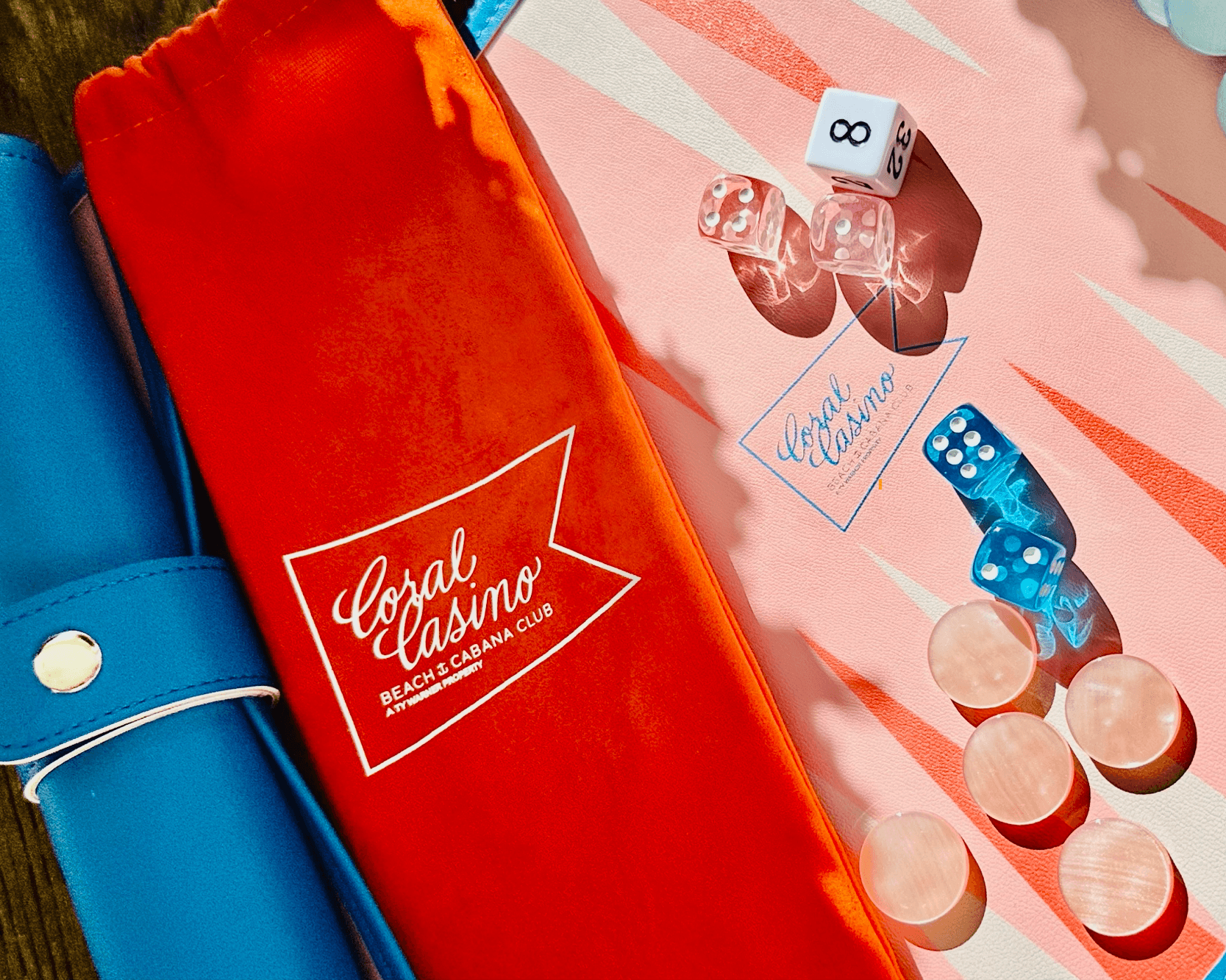 A close-up of a Coral Casino Beach & Cabana Club backgammon set — a red velvet pouch embroidered with the white Coral Casino crest on a coral-and-cream striped board with pink and blue dice and blush chips